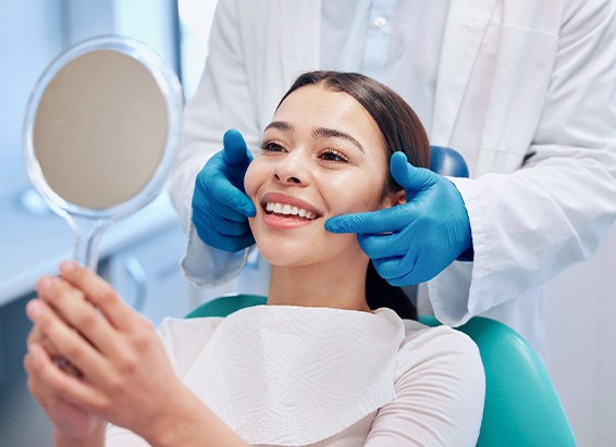 Woman in dental chair smiling at reflection with dentist behind her touching her cheeks