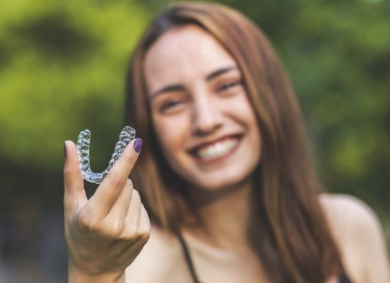 Smiling woman holding clear aligner for Invisalign in York outdoors